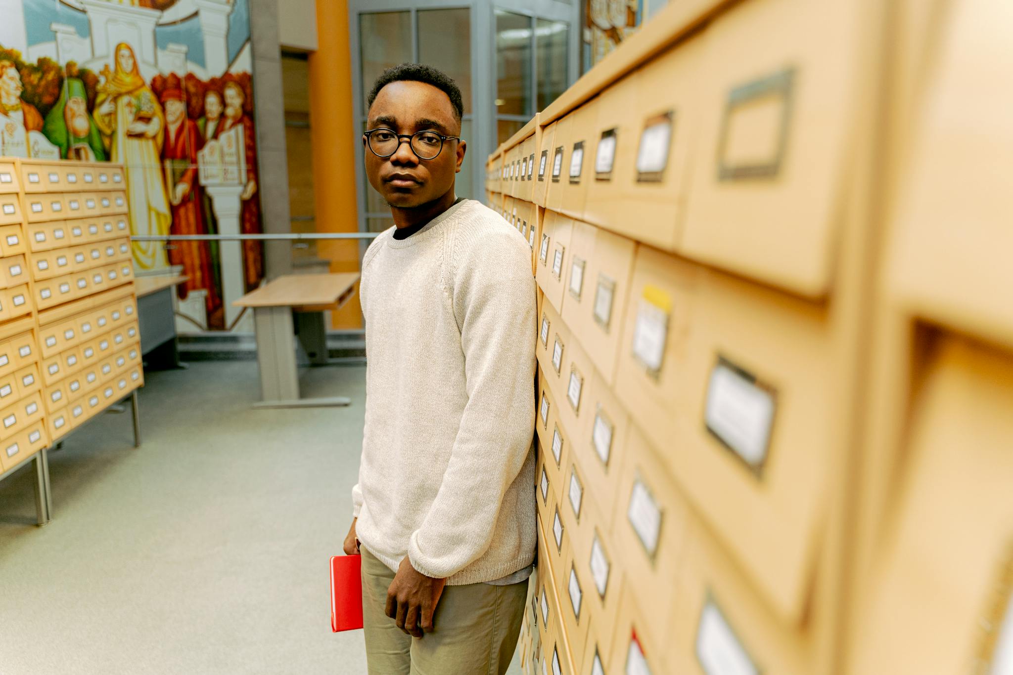 Young adult standing in a library corridor holding a red notebook. Surrounded by shelves and murals.